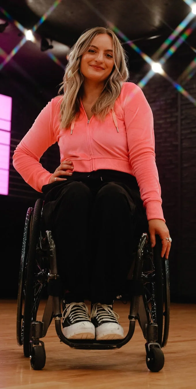 A photograph of Conner Lundius dancing in a wheelchair while wearing a coral zip-up hoodie and black pants and black and white sneakers. 