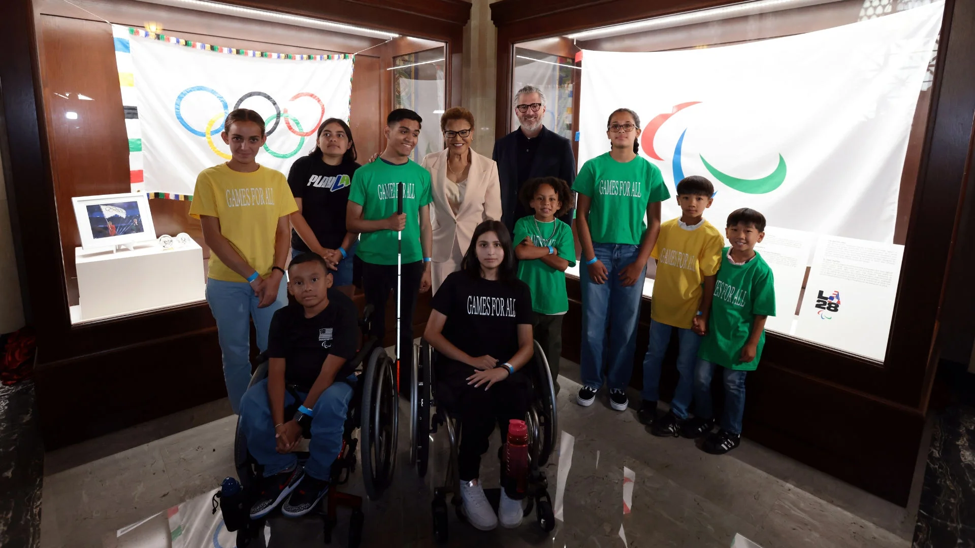 Photograph of Mayor Karen Bass, local city youth, and LA28 president and chairperson Casey Wasserman standing in front of the Olympic and Paralympic flag displays at Los Angeles City Hall. 