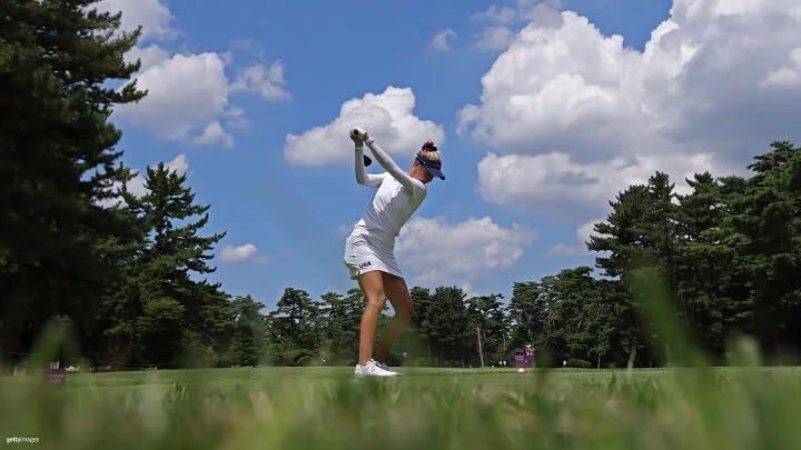 A female golfer is captured mid-swing on a bright, sunny day. She is dressed in a white long-sleeve shirt, a matching skirt, and a dark blue visor with the letters "USA" visible on her outfit. The backdrop features a clear blue sky with scattered white clouds and green trees framing the scene. The perspective is low to the ground, with green grass in the foreground.