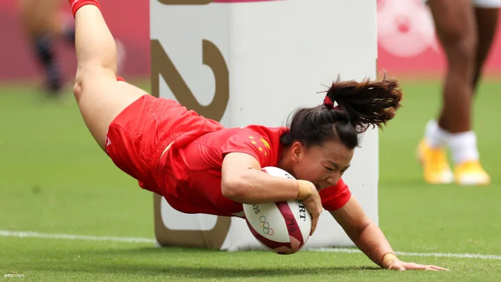 A female rugby player in a red uniform is diving to score a try, clutching the ball firmly. The player’s body is airborne with only one hand touching the grass near the try line. The background shows a blurred figure of another player and a padded post with markings.