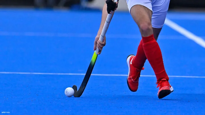 Una toma cercana de un jugador de hockey sobre césped en acción en un campo artificial azul brillante. El jugador lleva puestos calcetines rojos, zapatos rojos y pantalones cortos blancos. Sostiene un palo de hockey negro y amarillo neón, controlando una pelota blanca de hockey sobre el césped. El enfoque está en la parte inferior del cuerpo del jugador y el palo.