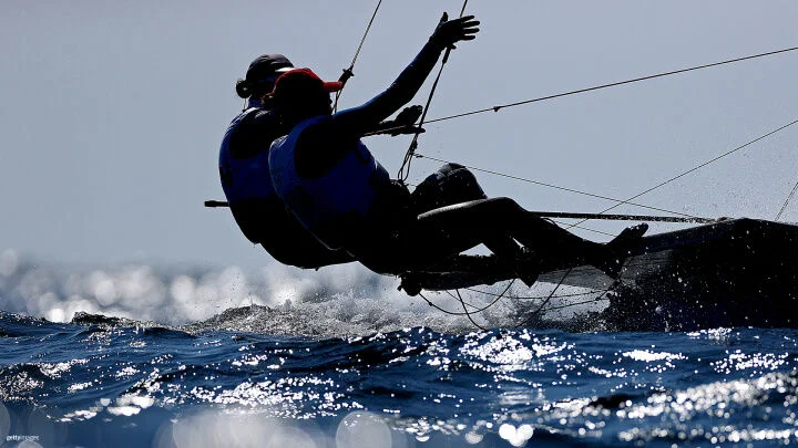 Two sailors in protective gear and life vests, one wearing a red cap, lean out over the edge of their boat, gripping the rigging lines. The bright sunlight reflects off the water, creating a dynamic scene with splashing waves.