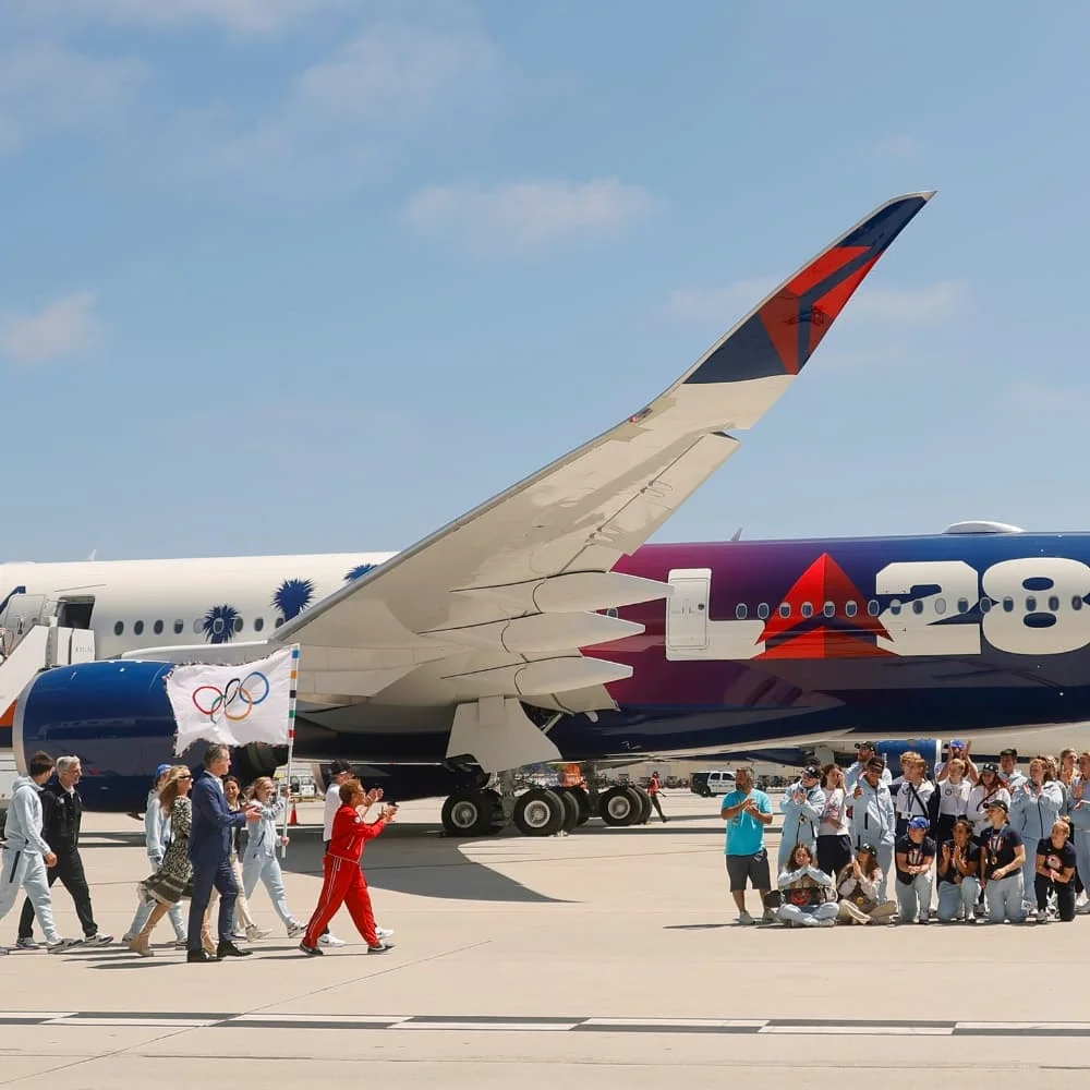 Photograph of California Governor Gavin Newsom holding the official Olympic flag while walking with LA Mayor Karen Bass, Team USA athletes and LA28 executives at the Delta Airlines hangar in Los Angeles.