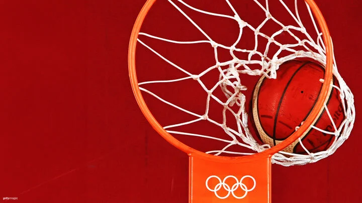 Se observa un balón de baloncesto atravesando el aro, capturado justo al pasar por la red. El aro y la red tienen un diseño estándar con el borde en color naranja y la red blanca. El tablero al que está fijado el aro presenta el logo de los anillos olímpicos, indicando que se trata de un evento de baloncesto olímpico. El fondo es de un color rojo liso.