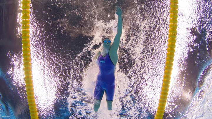 Una toma submarina captura a una nadadora Paralímpica de Team USA durante una competencia de estilo libre. Ella lleva puesto un traje de baño azul, gorro de natación y gafas. Su brazo izquierdo está extendido hacia arriba mientras se desliza por el agua, creando ondas y burbujas a su alrededor. Las piernas de la nadadora están amputadas debajo de las rodillas, y también le falta el brazo derecho. Los divisores amarillos visibles a ambos lados delinean su recorrido en la piscina.