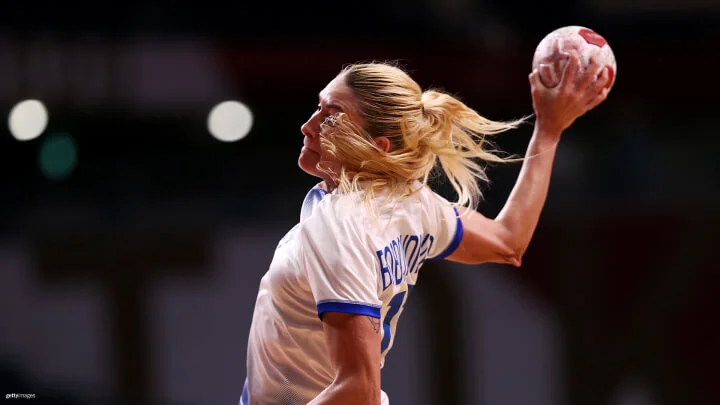 A medium shot captures a female handball player with blonde hair in a ponytail in action, about to throw the ball. She is wearing a white jersey with blue trim. The name on the back of her jersey is not fully visible.