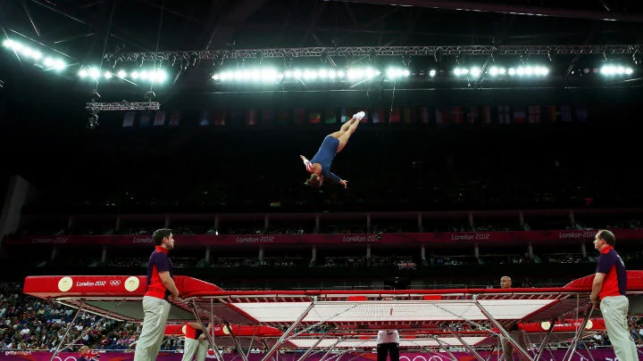 Una gimnasta en un maillot azul realiza una rutina de alto vuelo en un trampolín. Está suspendida en el aire, con su cuerpo completamente extendido en paralelo al suelo y los brazos extendidos hacia los lados. El trampolín está rodeado por asistentes en uniformes rojos y morados. El fondo muestra una gran arena llena de espectadores, banderas olímpicas y luces brillantes en el techo. Banderas nacionales de varios países cuelgan sobre la multitud.