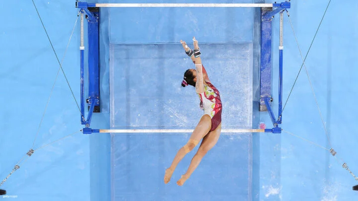 A female gymnast is captured mid-air during her routine on the uneven bars. She is seen from above, leaping between the bars with her body sideways, arms extended, and fingers pointed. She wears a red, green, and white leotard with intricate designs. Her hair is in a bun, and her wrists are wrapped with protective tape and grips. The background features a blue gymnastics mat with chalk marks.