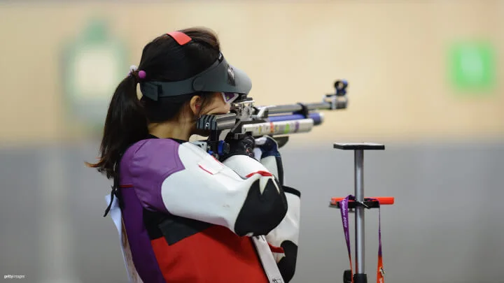 Una deportista está apuntando con un rifle de aire comprimido hacia un blanco en un campo de tiro cubierto. Lleva puesta una chaqueta de tiro en colores morado, rojo y blanco, guantes, protección ocular y una visera, con el cabello recogido en una cola. El rifle está apoyado en su hombro en posición de disparo.