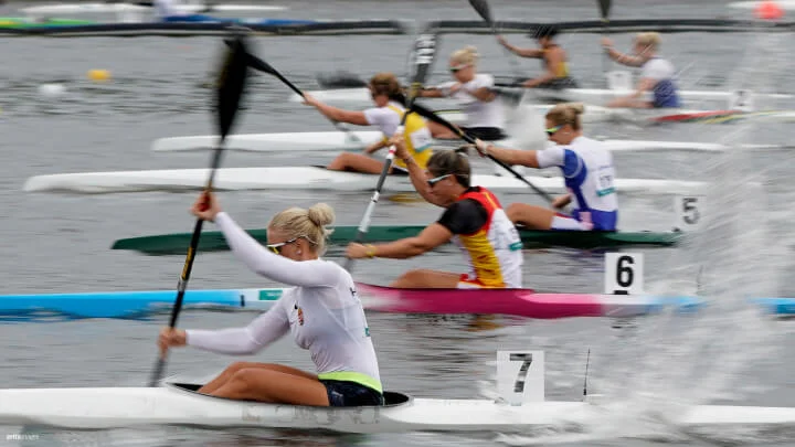 Una toma dinámica de una carrera de canoas en un cuerpo de agua tranquilo con varias atletas femeninas remando enérgicamente. Las canoas están agrupadas con marcadores de carril visibles numerados del 1 al 7 de derecha a izquierda. La atleta en primer plano, vestida con una camiseta blanca de manga larga y gafas de sol, está en el kayak número 7. Los atletas están en pleno remo, generando salpicaduras al remar vigorosamente en el agua.