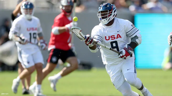 A male lacrosse player wearing a white uniform with "USA" and the number 9 on the front is in action on the field. The player is holding a lacrosse stick and appears to be tracking or about to catch a yellow lacrosse ball in mid-air. The player is also wearing a white helmet with a blue visor, white gloves, and arm guards. In the background, another player in a white uniform and one in a red uniform are visible, with the game taking place on a green field.