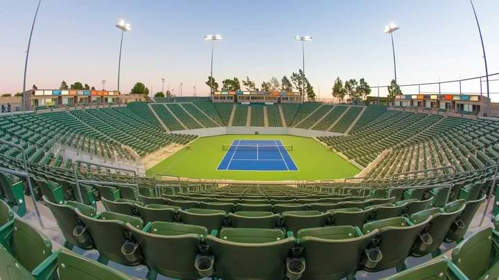 Photograph of a tennis court showing the entire seating area in an oval shape with green seats and stadium lights surrounding the court. The tennis court has a net stretched across a blue court which is bordered by green flooring. In the background there are a few trees and blue sky.