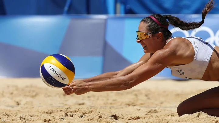 A beach volleyball player wearing a white sports bra and sunglasses dives in the sand to dig the ball. She has a focused expression, and her hair is tied back in a ponytail. The ball is yellow, blue, and white, marked with "Tokyo 2020." The background features a blurred view of the Olympic rings and a blue barrier.