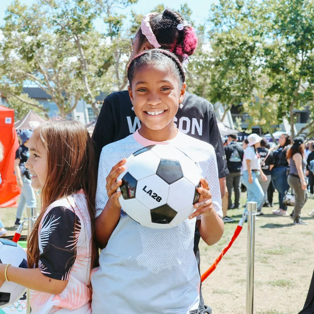 Photo of a young girl in a white tshirt smiling and holding up a soccer ball with LA28 prined on the ball