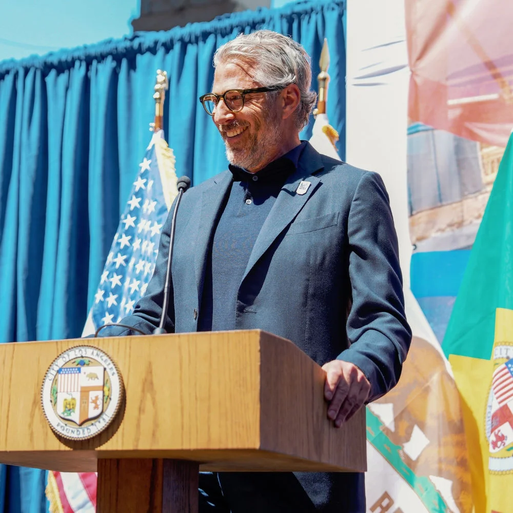 Photograph of LA28 chairperson and president Casey Wasserman standing at a podium with a microphone during the Olympic and Paralympic Flag Installation ceremony at Los Angeles City Hall.