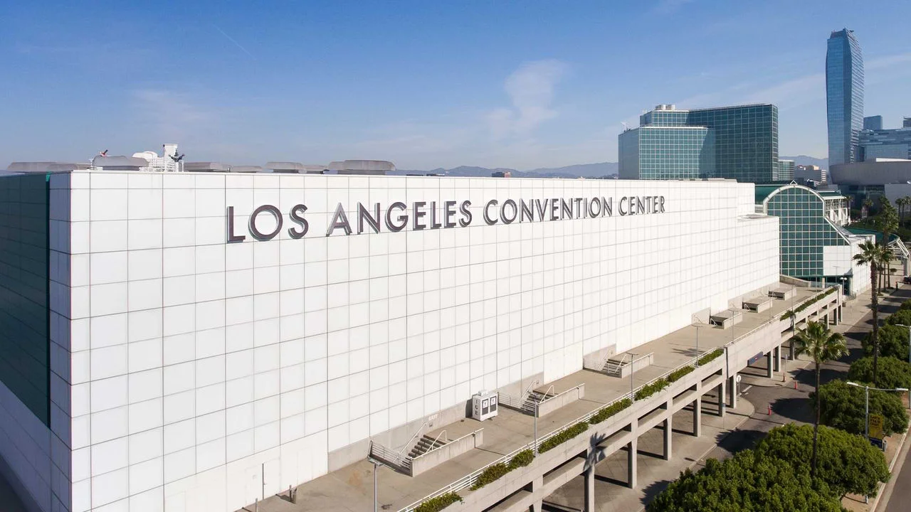 Photograph of the Los Angeles Convention Center in Downtown Los Angeles. It is a large white rectangular building with the words  "Los Angeles Convention Center" in large silver letters. The building design has multiple small squares covering the entire thing and the background is an LA skyline.