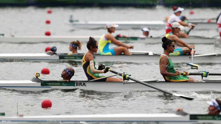 Varios equipos de remo están participando en una competencia en una superficie serena de agua, donde las boyas rojas indican las rutas. Destacado en primer plano, se observa claramente un equipo de Brasil, compuesto por dos remeros vestidos con uniformes en verde, negro y amarillo, junto a un timonel que mira en dirección opuesta. Ambos remadores agarran sus remos y los mueven hacia atrás al unísono. Uno de los remeros tiene el brazo vendado, y ambos llevan gafas de sol. Otros equipos son visibles en el fondo, con remeros en uniformes de diferentes colores de distintos países.