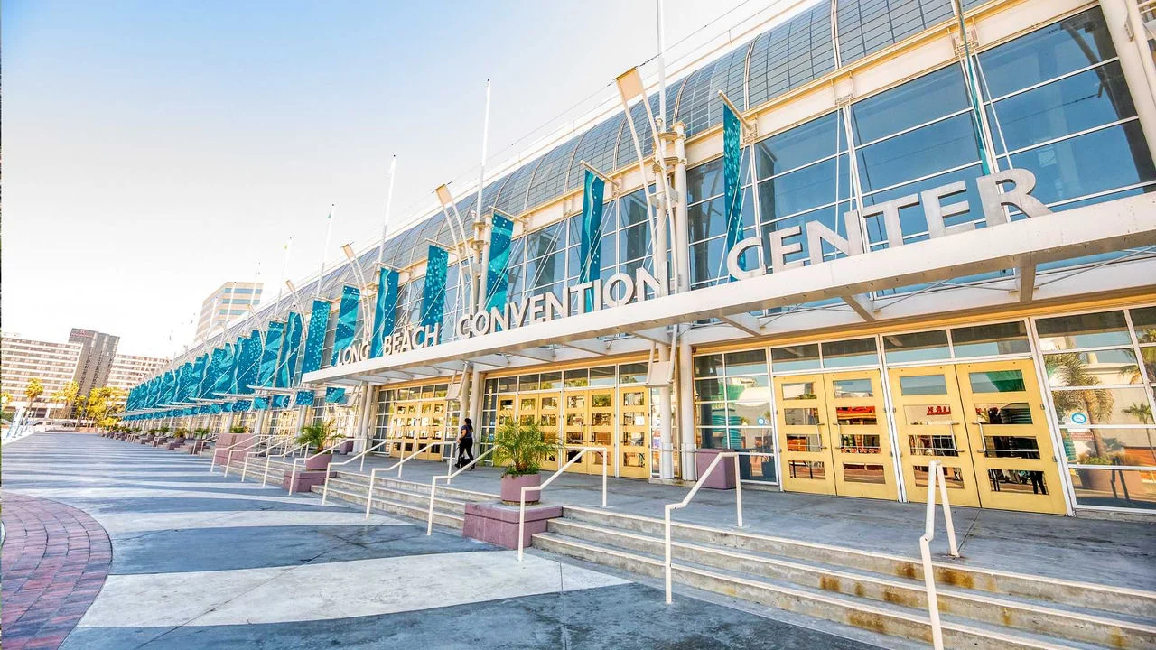 Photograph of the Long Beach Convention Center, a long rectangular building with yellow doors, and an arched glass front with concrete flooring outside, steps and white hand rails and blue flags hanging from the front. The words 'Long Beach Convention Center' are above the entrance in large silver letters.