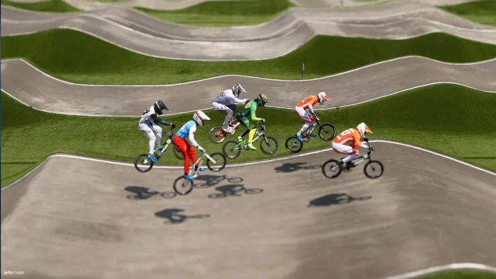 A group of BMX racers is captured mid-race on a track featuring numerous undulating hills. Each rider is airborne, their bikes lifted off the ground as they navigate the course. The racers wear colorful racing outfits, helmets, and protective gear. The track is a mix of green artificial turf and light grey racing paths. The shadows of the riders and their bikes are visible on the track below, emphasizing the height of their jumps.