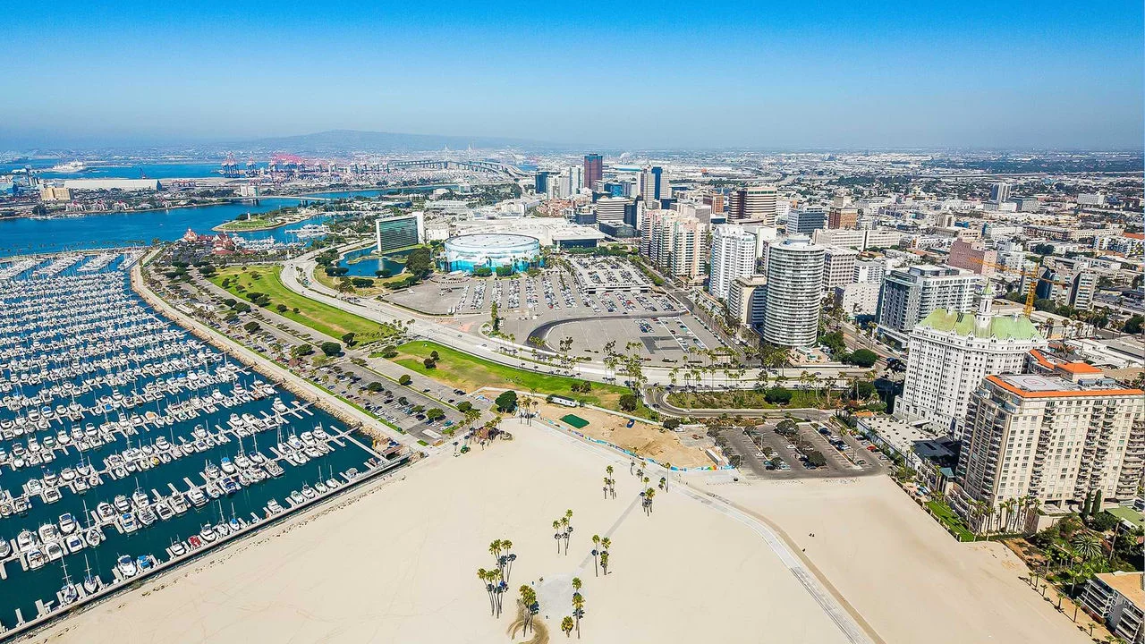 Fotografía aérea del paseo marítimo en Long Beach con un puerto deportivo lleno de botes a la izquierda, una zona de playa arenosa en el centro frontal, un estacionamiento en el centro y numerosos edificios y rascacielos a la derecha, con un cielo azul y vistas de la ciudad en el fondo.