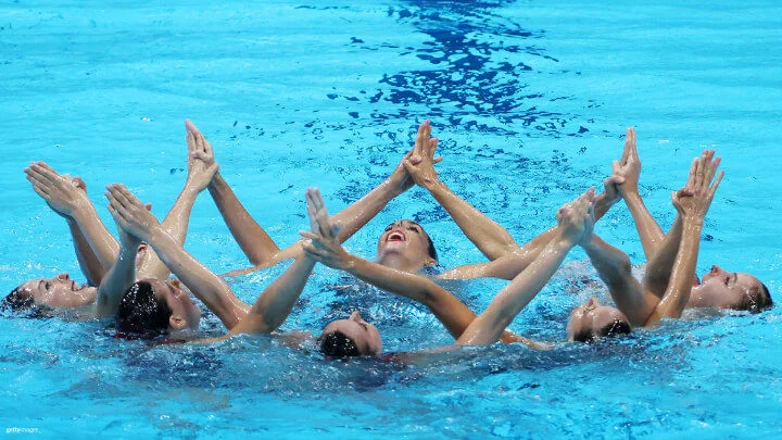 A group of artistic swimmers is captured performing a routine in a bright blue pool. They are arranged in a circle, with their heads above the water and their arms extended upwards, touching at the fingertips to form a star-like shape. The swimmers are smiling and appear focused as they execute their choreography. 