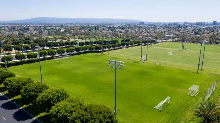 Fotografía desde arriba de un campo grande de fútbol con cuadros de objetivo final en cada extremo y césped verde brillante, rodeado por arboles verdes además de luces de estadio. En el fondo hay un paisaje urbano que muestra casas y edificios, cielo azul y las montañas en la distancia. 