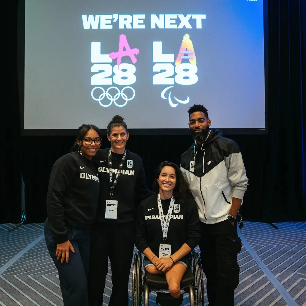 Group photo featuring Athlete Fellows who were Olympians and Paralympian athletes now full-time LA28 employees standing in front of a projector screen that shows the LA28 dual emblems with the words 'We're Next'