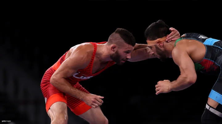 Two muscular male wrestlers are engaged in a match against a dark background, bent forward and gripping each other's heads. The wrestler on the left is wearing a red singlet with "USA" printed on it, while the wrestler on the right is wearing a black and blue singlet with orange athletic tape on his head.