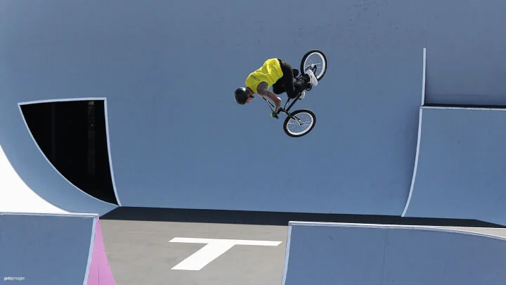 Un ciclista de BMX está realizando una acrobacia aérea muy por encima de las rampas en un skatepark. Viste una camiseta amarilla brillante, pantalones negros y un casco negro, y se le captura en el aire, sentado en la bicicleta de BMX con el cuerpo inclinado hacia adelante. Las rampas y superficies del skatepark son de color azul claro, gris y blanco.