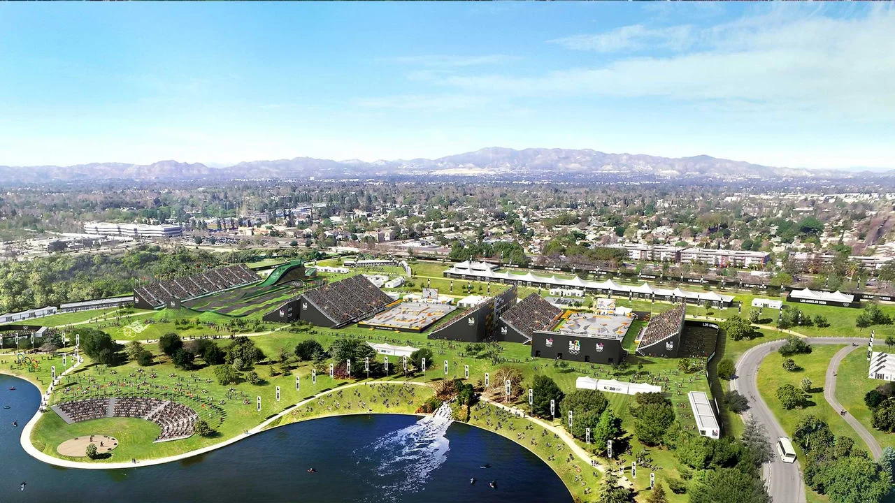 Rendering of the Sepulveda Basin Recreation Area set up for the Los Angeles 2028 Olympic and Paralympic Games. The area features multiple sports venues with grandstands filled with spectators. The venues include a BMX track with an elevated ramp and a skate park with various ramps and obstacles. Surrounding the sports venues are green spaces. A large lake with a small waterfall is visible in the foreground. The background showcases urban landscape with mountains. "Los Angeles 2028" banners and Olympic-themed decorations are visible throughout the area.