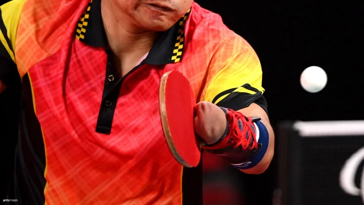 A Paralympic table tennis player, wearing a vibrant red and yellow shirt, is in action as he hits the ball. The athlete, who is a hand amputee, has the paddle strapped to his residual limb with a red and blue strap. The ball is visible in mid-air, heading towards the player against a dark background.