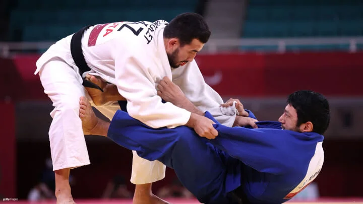 Two judokas are engaged in a judo match. The judoka on the left is wearing a white gi and has a grip on the collar and sleeve of the judoka on the right, who is wearing a blue gi. The judoka in blue is on the ground, using his right leg to push against the hip and leg of the judoka in white while gripping the white gi with his hands. 