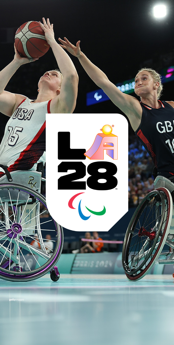 Photograph of two female wheelchair basketball players reaching for the ball while wearing navy blue and white basketball uniforms with the LA28 Determination Paralympic A emblem overlayed on top.