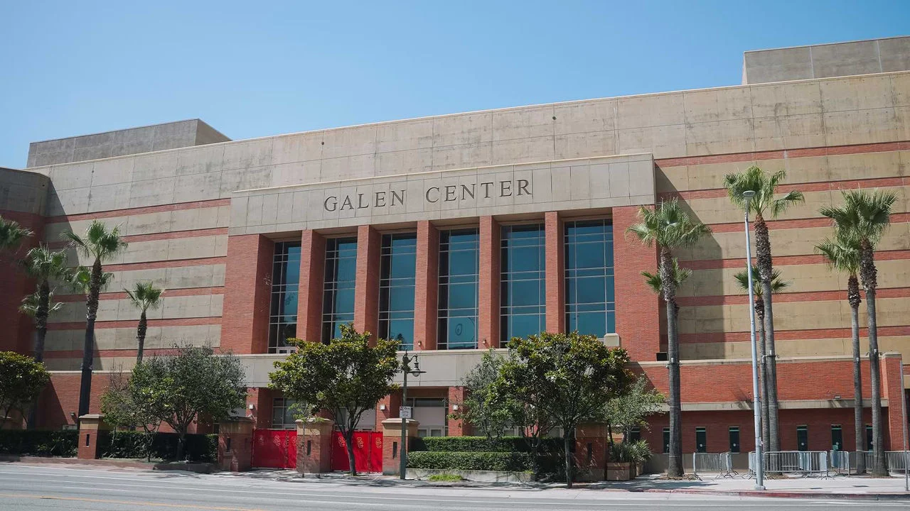 Photograph of Galen Center, a tall rectangular building with red and tan colored bricks and six tall rectangular windows in the center, with numerous palm trees and other foliage near the front of the building and a blue sky in the background.