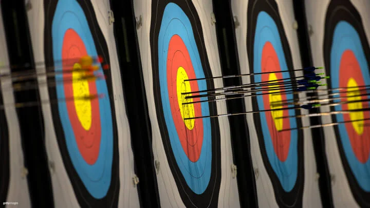 A row of archery targets is shown with multiple arrows embedded in them. The targets feature the standard concentric rings colored black, blue, red, and yellow, with the arrows clustered mainly in the yellow bullseye area, indicating accurate shots.