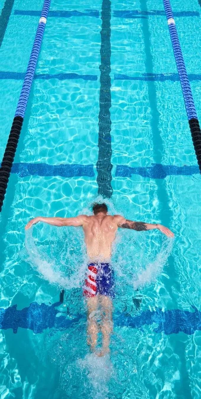 Fotografía demostrando un hombre que tiene una manga de tatuajes en su brazo derecho, nadando en una piscina de color aguamarina azul mientras lleva puesto un par de pantalones cortos con un estampado de la bandera estadunidense. 