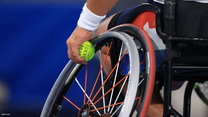 Una imagen cercana muestra un jugador de tenis en silla de ruedas sosteniendo una pelota de tenis. La mano de el atleta esta agarrando una pelota de tenis amarilla con la frase "Tokio 2020" impreso en él contra la rueda de su silla de rueda deportiva. La silla de ruedas tiene un marco negro, llantas rojas, radios rojas y blancas en las ruedas, y la bandera francesa en los protectores laterales. El atleta lleva una muñequera blanca. 