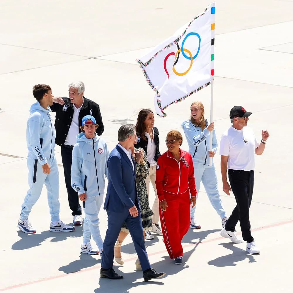 Fotografía en primer plano de un atleta del Team USA izando la bandera olímpica oficial mientras un grupo en el que se encuentran la alcaldesa de Los Ángeles, Karen Bass, el gobernador de California, Gavin Newsom, la esposa del gobernador Newsom,Jennifer Siebel Newsom, y ejecutivos de LA28 caminan por el hangar de Delta Airlines en Los Ángeles. 