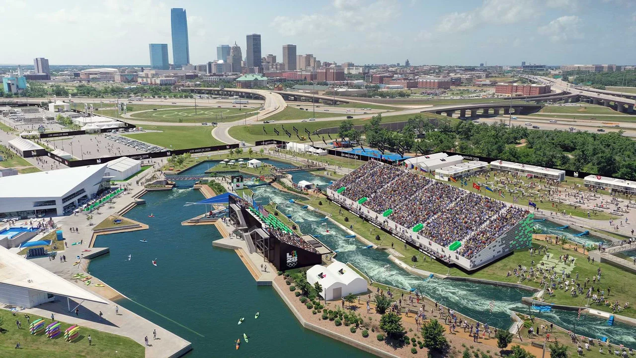 A digital rendering shows the Whitewater Center, also known as the Riversport OKC. The image includes a large water course with kayakers navigating rapids. There are packed grandstands full of spectators watching the event. The surrounding area has several buildings, tents, and pathways branded with the LA28 logo. In the background, the skyline of Oklahoma City with tall buildings and highways is visible. The event banners read "Los Angeles 2028," indicating it is an Olympic venue.