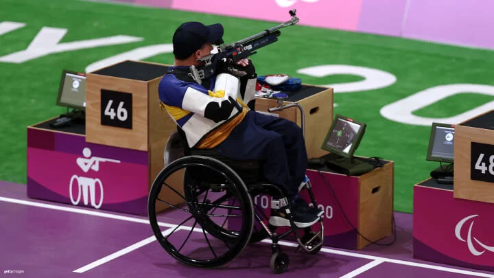 A Paralympic athlete competes in a shooting event at the Tokyo 2020 Paralympic Games. The athlete is seated in a wheelchair, wearing a blue, white, and yellow shooting jacket and a navy cap. They are aiming an air rifle at the target in the distance. The shooting range has designated lanes marked with numbers, and the athlete is in lane 47. Electronic scoring monitors are positioned on tables beside each lane. The event's logo, featuring a stylized figure in a wheelchair aiming a rifle, and the Paralympic Agitos are visible on the lane markers.