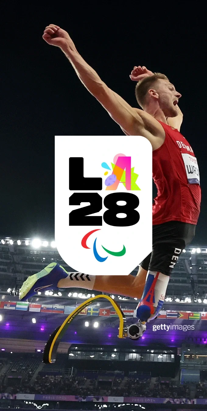 A photograph of a Paralympian long jumping at the Paris 2024 Olympics with stadium lights and seating in the background. The LA28 Boldness Paralympic 'A' emblem is overlayed in the middle of the photograph. 