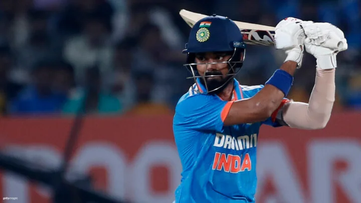 An Indian cricketer in action is captured in this image. He is wearing the blue team jersey with "INDIA" printed on it in orange, and a dark blue helmet with the Indian flag and emblem. He is seen holding the bat raised behind his head. The background is a blur of the stadium crowd.