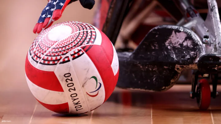 A close-up image of a red and white rugby ball on a wooden court, with "Tokyo 2020" and the Paralympic logo printed on it. The ball is about to be picked up by a gloved hand wearing a glove with an American flag design. Part of a wheelchair, with scuff marks and red wheels, is visible next to the ball. The focus is on the ball and the hand, with the wheelchair slightly blurred in the background.