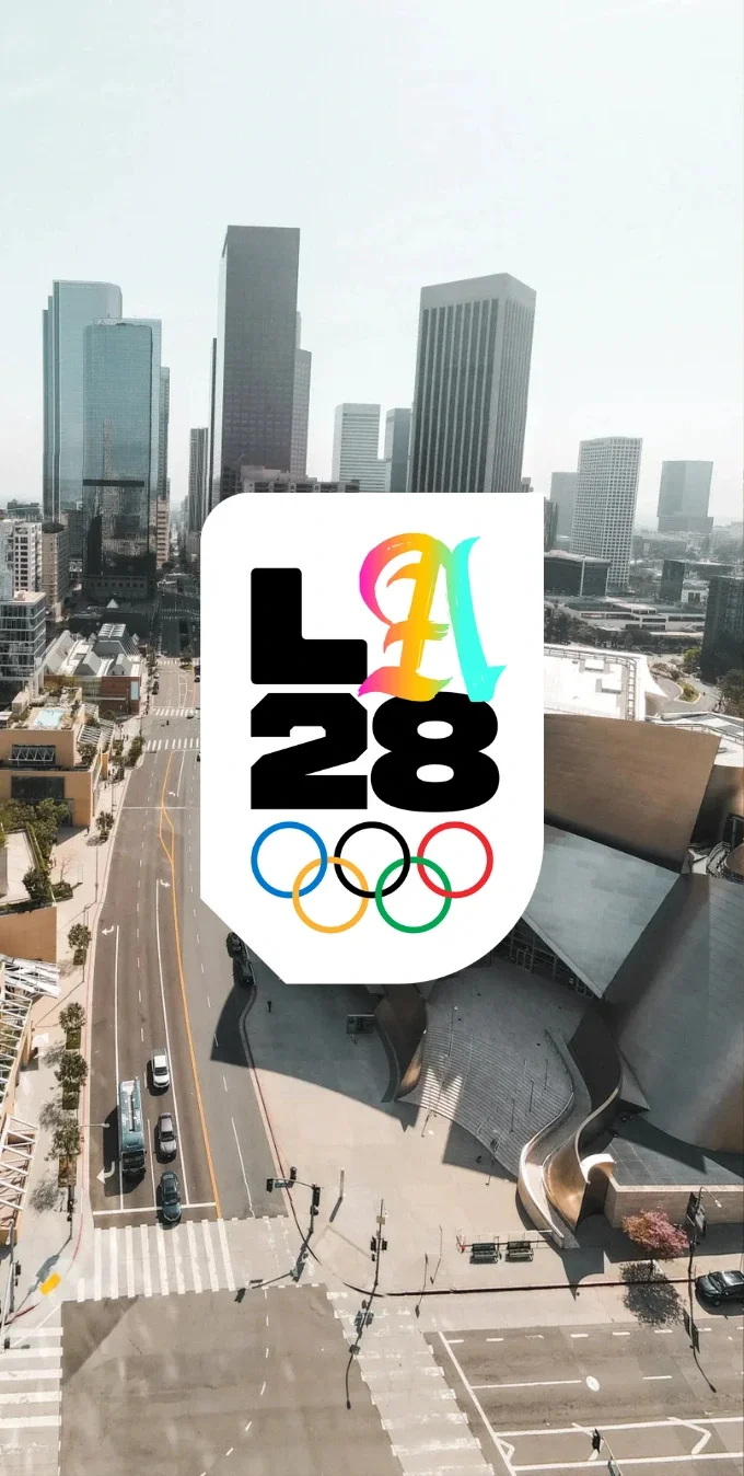 A photograph of the downtown Los Angeles skyline with the Walt Disney Concert Hall in the foreground. The LA28 Gradient Script Olympic A emblem is overlayed on top of the photograph.