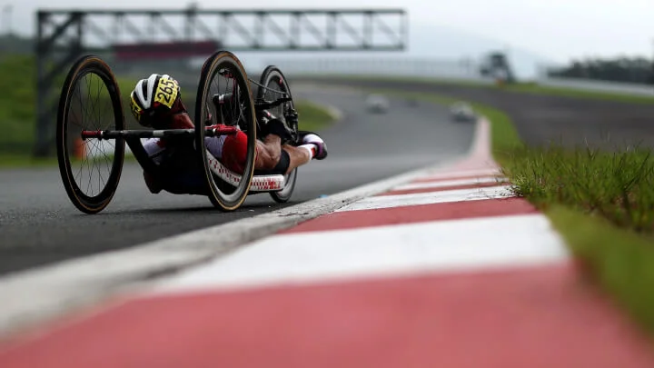 Una vista desde un ángulo bajo de un ciclista de mano compitiendo en una pista. El atleta, con un casco blanco y un dorsal amarillo con el número 265, está recostado en su handbike, impulsándose hacia adelante con los brazos. La pista tiene marcas rojas y blancas en el bordillo, y el fondo borroso muestra la curva de la pista hacia adelante.