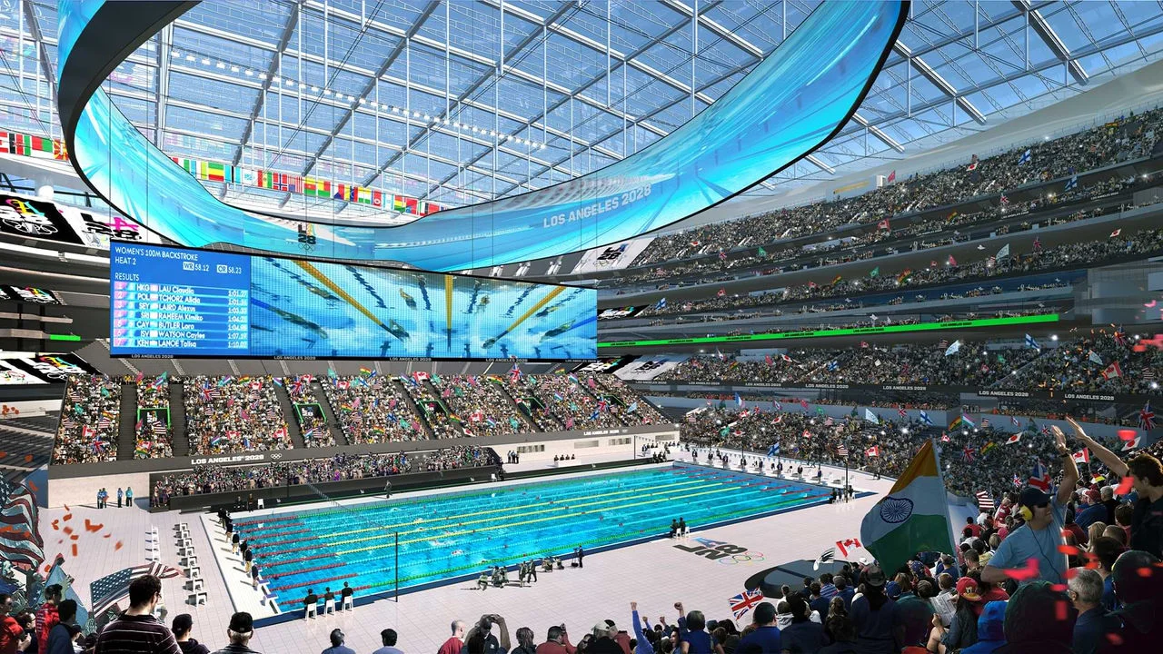 Rendering of the 2028 Olympic and Paralympic Stadium, also known as SoFi Stadium. The modern indoor venue is filled with fans waving flags from different countries. The focus is on the swimming pool where athletes are racing. Large screens above the pool show the Women's 100m Backstroke event and live footage of the swimmers. The stadium has a clear roof and many levels of seating, all filled with spectators.