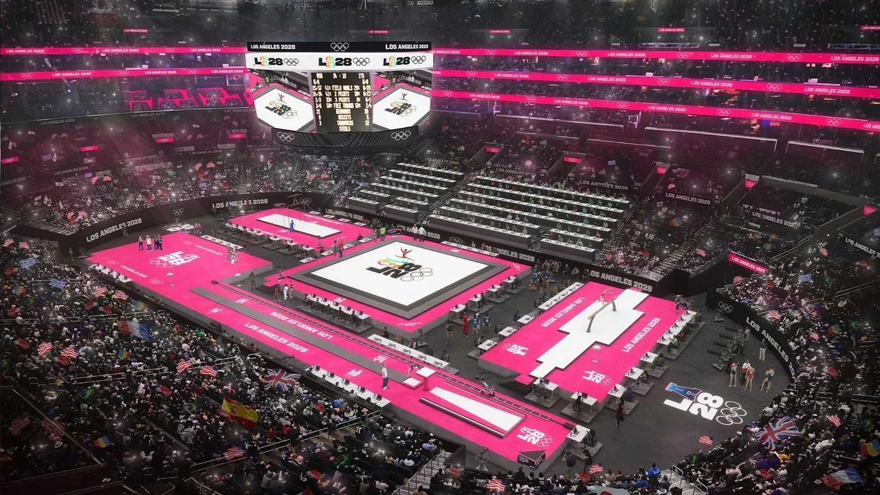 Rendering of the Los Angeles 2028 Olympic and Paralympic Games Arena, also known as the Crypto.com Arena. The arena is filled with spectators, with the seating areas displaying vibrant pink signage indicating "LOS ANGELES 2028" in white text. Multiple gymnastics apparatuses are set up on the floor, including the balance beam, uneven bars, and floor exercise area, all with pink flooring. Athletes and officials are visible near the apparatuses. Large digital screens show the event's logo, scores, and event information. The audience waves various international flags.