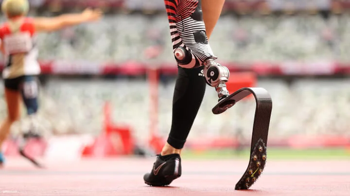 Una imagen cercana de la cuchilla protésica izquierda de un velocista en una pista. La prótesis tiene un diseño de rayas negras, rojas y blancas en el acoplador de la pierna, la cuchilla está hecha de fibra de carbono, y el atleta lleva zapatos para correr negros y una manga negra en la pierna derecha. En el fondo borroso, se puede ver a otro atleta con una pierna protésica.