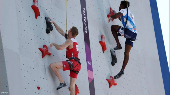 Dos escaladores compiten ascendiendo una pared vertical en un evento de escalada deportiva. El escalador de la izquierda lleva un uniforme rojo y blanco con "Austria" impreso en él. El escalador de la derecha lleva un uniforme azul y blanco. Ambos están agarrando presas rojas y negras y están asegurados con arneses y cuerdas. El logo "Tokio 2020" está destacado en el centro de la pared.