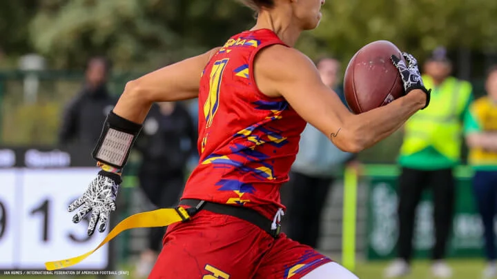 Una imagen cercana muestra a un jugador de flag football en acción. El jugador viste una camiseta roja sin mangas con diseños geométricos en amarillo y azul, guantes blancos con patrones, y una muñequera negra con un cuaderno táctico atado a su muñeca izquierda. Sostiene un balón de fútbol americano en su mano derecha mientras corre. Una bandera amarilla cuelga de su cintura, indicando un juego de flag football. En el fondo se ven espectadores y oficiales borrosos.